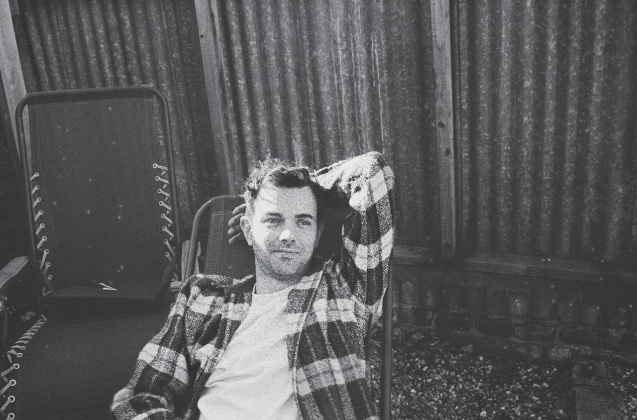 Black and white press photo of Tom Iansek of #1 Dads wearing a plaid jacket and reclining outdoors by a corrugated fence.